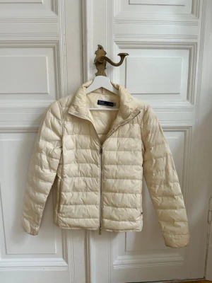 Ljusbeige dunjacka med dragkedja - Snygg ljusbeige dunjacka med quiltat mönster och hög krage. Jackan har dragkedja framtill och två sidofickor. Perfekt för kalla vinterdagar och ger en clean och stilren look. Materialet är lätt och värmande.