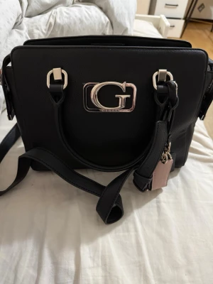 Svart handväska från Guess - Snygg svart handväska från Guess med stort G-logotyp i silver på framsidan. Väskan har både handtag och en avtagbar axelrem samt detaljer i metall. Tillverkad i slätt konstläder och har en klassisk, fyrkantig form.