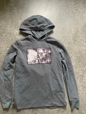 Grå hoodie med boxningsmotiv - Snygg grå hoodie med huva och ett stort svartvitt tryck på bröstet som visar en klassisk boxningsscen. Hoodien har långa ärmar och ribbade muddar. Perfekt för dig som gillar sportiga och unika plagg med coola motiv. Märker jag skrev är inte märket på hoodien 