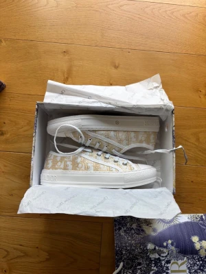 Beige Dior sneakers med monogram - Säljer ett par snygga Dior sneakers i beige och vitt med klassiskt Dior-monogram. Skorna har vita snören, gummisula och lågt skaft. Perfekta för dig som vill ha en exklusiv touch på din outfit. Kommer med originalkartong och dustbag.