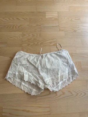 Vita broderade shorts med volangkant - Säljer ett par vita shorts i bomull med broderade detaljer och volangkant. De har elastisk midja och ett luftigt, löst passform som passar perfekt till varma dagar. Supersöta och lätta att matcha med olika toppar.