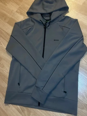 Grå hoodie med dragkedja från BOSS - Snygg grå hoodie från BOSS med dragkedja framtill och justerbar huva med svarta snören. Diskret BOSS-logga på bröstet och två fickor med dragkedja. Perfekt för en avslappnad och stilren look.