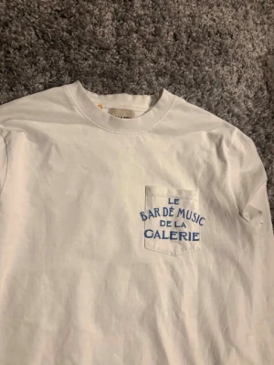Vit långärmad topp Gallery Dept - Vit långärmad topp från Gallery Dept med blå texttryck på bröstficka, rygg och ärm. Klassisk rund halsringning och relaxed passform. Perfekt för dig som gillar streetwear och vill sticka ut med coola detaljer.