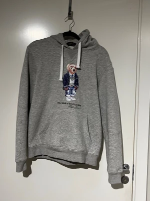 Grå hoodie Polo Ralph Lauren - Snygg grå hoodie från Polo Ralph Lauren med ikoniska björnmotivet på bröstet. Hoodien har vita dragsnören, känguruficka och ribbade muddar. Tillverkad i mjukt bomullsmaterial som är perfekt för chill dagar. Klassisk och stilren design som alltid funkar.
