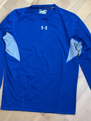 Blå långärmad träningströja Under Armour - Snygg blå långärmad träningströja från Under Armour med gråa paneler vid sidorna. Perfekt för träning eller löpning, tillverkad i ett stretchigt och snabbtorkande material. Diskret logga framtill och text på ryggen.