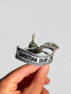 Christian Dior tygarmband - Snyggt tygarmband från Christian Dior med texten 'J'adior' i svart på vit bakgrund. Armbandet har flätade band och tofsar i ändarna, vilket ger en cool och trendig look. Perfekt accessoar för att lyfta din outfit.