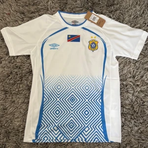 Vit och blå fotbollströja Umbro - Säljer en vit fotbollströja från Umbro med blå detaljer och grafiskt mönster nedtill. Tröjan har tryckt flagga och klubbmärke på bröstet samt rund halsringning med blå kant. Perfekt för dig som gillar sportig stil.