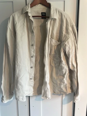 Ljusbeige jeansjacka från Zara - Stilren ljusbeige jeansjacka från Zara med klassisk krage, knappstängning och en bröstficka. Jackan har en avslappnad passform och är tillverkad i ett mjukt jeanstyg som ger en casual vibe. Perfekt att slänga över en hoodie eller t-shirt.