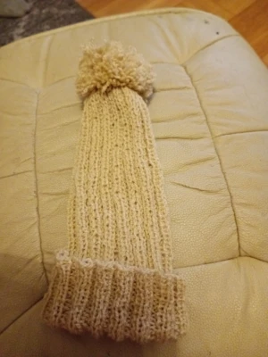 Beige stickad mössa med tofs - Säljer en beige stickad mössa med ribbad struktur och en stor fluffig tofs på toppen. Mössan är lång i modellen och har en vikt kant nedtill. Perfekt för dig som vill ha en enkel men snygg accessoar till hösten eller vintern.