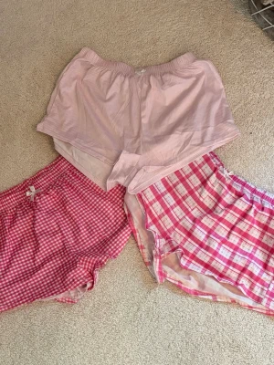 3-pack rosa pyjamasshorts - Säljer ett 3-pack pyjamasshorts i olika rosa mönster: ett par med smala ränder, ett par med små rutor och ett par med större rutmönster. Alla har elastisk midja och mjukt material, perfekta för mysiga kvällar hemma. Skriv pm vid funderingar!