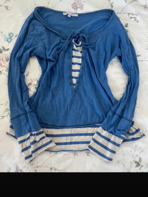 Blå långärmad topp från La Petite Francaise - Blå långärmad topp från La Petite Francaise med snörning framtill och v-ringad öppning. Toppen har vita och blå ränder på ärmslut och nederkant som ger en cool kontrast. Tillverkad i mjuk bomull, perfekt för en avslappnad look. Små hål (se bild)