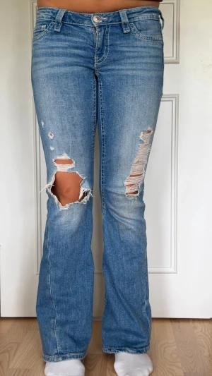 Blå bootcut jeans - Säljer ett par ljusblå bootcut jeans med slitningar och hål på båda benen. Jeansen har klassiska fickor. Bakfickorna har lock med knappdetaljer vilket är trendigt nu. Perfekta för en avslappnad och trendig look.