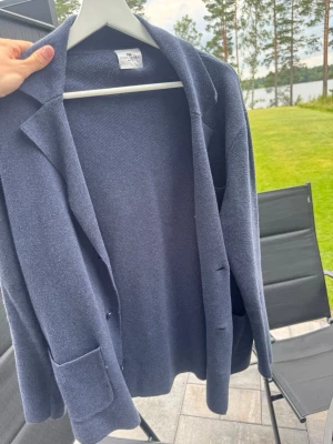 Mörkblå kavaj med fickor - Stilren mörkblå kavaj med klassisk krage och två stora fickor framtill. Kavajen har en rak passform och är tillverkad i ett mjukt, stickat material som ger en avslappnad men ändå dressad look. Perfekt att slänga över en t-shirt eller skjorta.