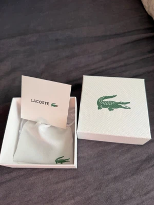 Lacoste silverfärgat armband - Snyggt armband från Lacoste i silverfärgad metall med rektangulära länkar. Kommer i original ask och påse med klassiska Lacoste-loggan. Perfekt accessoar för dig som gillar stilrena detaljer och vill ha något som sticker ut lite extra.