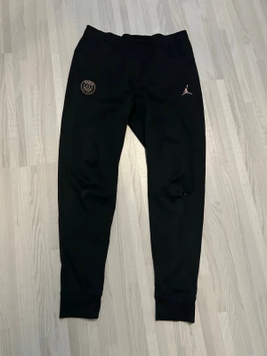 Svarta PSG x Jordan träningsbyxor - Säljer ett par svarta träningsbyxor med Paris Saint-Germain och Jordan-logga i guld på framsidan. Byxorna har resår i midjan och vid bensluten, samt smal passform. Perfekta för träning eller chill.