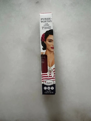 theBalm Purse-Worthy Lip Gloss Wristlet - Snyggt läppglans från theBalm i nyansen Wristlet. Färgen är en djup röd med glansig finish. Kommer i en smidig förpackning med retroinspirerad design och illustration. Innehåller 7 ml och är både vegansk och cruelty free. Oöppnad förpackning.