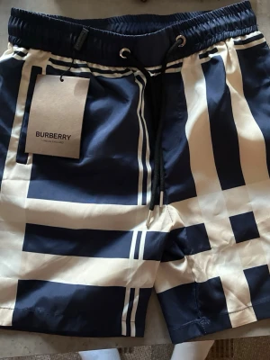 Marinblå-vita badbyxor Burberry - Snygga badbyxor från Burberry i marinblått och vitt med klassiskt rutmönster. De har resår i midjan, dragsko och sidofickor. Materialet är lätt och snabbtorkande, perfekt för stranden eller poolen. Stilsäker design med exklusiv känsla.