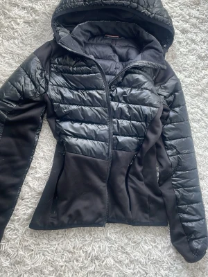 Svart ridjacka med huva hööks - Säljer en svart tunn ridjacka med huva och dragkedja framtill. Jackan har quiltade partier i glansigt material på bröst, ärmar och huva samt mjukare tyg på sidor och rygg. Två fickor med dragkedja och figurnära passform. 