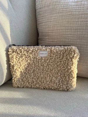 Beige Teddy necessär/fodral från Emelo - Säljer en beige, fluffig necessär från Emelo med mjuk teddystruktur. Väskan har en rektangulär form och stängs med dragkedja upptill. Perfekt för att förvara småsaker eller smink. Enkel och stilren design med en liten logotyp framtill. Mått ca 26x17cm