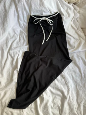 Svart baddräkt med vit knytning - Snygg svart baddräkt med vit knytning framtill i midjan. Klassisk modell med hög skärning och enkel design. Tillverkad i stretchigt material som passar perfekt för stranden eller poolen.