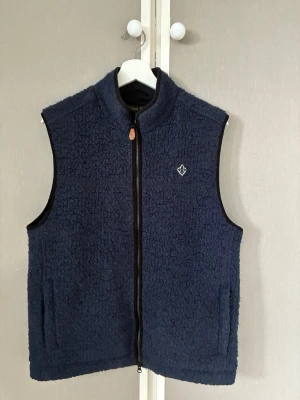 Morris väst blå storlek M - Mysig mörkblå väst i teddyfleece från Morris med dragkedja framtill och två fickor. Västen har en liten broderad logga på bröstet och ribbad krage. Perfekt för lager-på-lager och ger en cool, avslappnad stil.