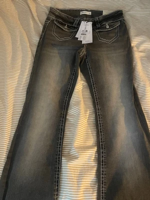 Grå bootcut jeans med kontrastsömmar - Nya snygga grå bootcut jeans med markerade vita kontrastsömmar och dekorativa bakfickor. Jeansen har låg midja och klassisk femficksdesign. Perfekta för dig som gillar en avslappnad men trendig stil.
