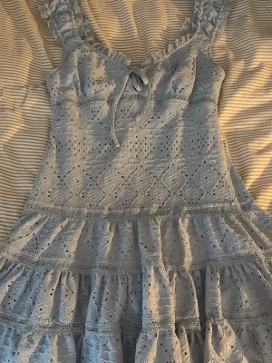 Ljusblå spetsklänning med volanger - Söt ljusblå klänning med broderad spets, volanger och knyt i halsen. Klänningen har breda axelband med volangdetaljer och ett romantiskt mönster. Perfekt för sommarens alla tillfällen.