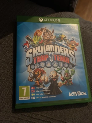 Skylanders Trap Team Xbox One spel - Skylanders Trap Team till Xbox One i original grön plastfodral. Omslaget har färgstarka illustrationer av olika Skylanders-karaktärer och spelets logga. Spelet är PEGI 7 och har stöd för svenska, norska, danska och finska.
