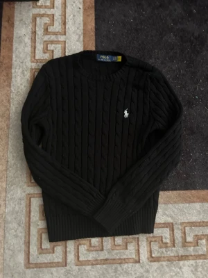 Svart kabelstickad tröja Polo Ralph Lauren - Kabelstickad svart tröja från Polo Ralph Lauren med rund halsringning och den klassiska vita logotypen broderad på bröstet. Tröjan har långa ärmar och ribbade muddar vid ärmslut och nederkant. Perfekt för en stilren och tidlös look.