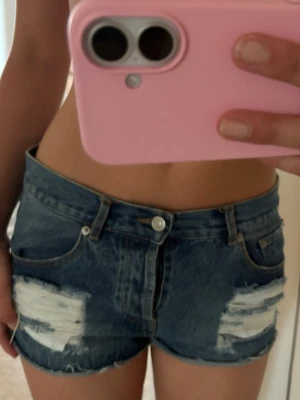 Blå jeansshorts med slitningar - Säljer ett par blå jeansshorts med råa kanter och slitna detaljer framtill. Shortsen har klassisk femficksdesign och knappgylf. Perfekta för sommaren och ger en avslappnad streetstyle-look.