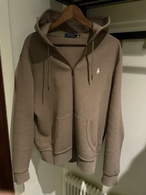 Full zip Polo Ralph Lauren - Snygg beige hoodie från Polo Ralph Lauren med dragkedja, huva med dragsko och kängurufickor. Klassisk logga broderad i vitt på bröstet. Tillverkad i mjuk bomullsblandning som känns skön mot huden. Användts ett handfull antal gånger, säljes då den inte riktigt passade mig. Är L men skulle säga att det är en stor M alternativt liten L.