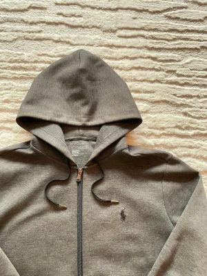 Grå zip hoodie från Polo Ralph Lauren - Snygg grå hoodie med dragkedja från Polo Ralph Lauren. Klassisk design med huva och dragsnören, samt den ikoniska loggan broderad på bröstet. Tillverkad i mjukt material som känns skönt mot huden. Perfekt för en avslappnad och stilren look. Inga skavanker. Längd 66cm. Bredd 55cm