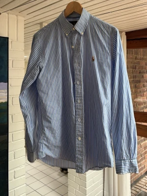 Ralph Lauren skjorta - Klassisk blå och vit randig skjorta med button-down krage och långa ärmar som är aldrig tvättad och inprincip ny. Skjortan har knappar framtill och en liten broderad logga på bröstet. Tillverkad i bomull och passar perfekt till en avslappnad men ändå stilig look.