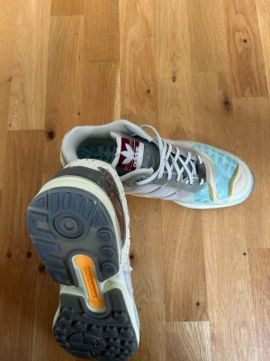 Adidas höga sneakers i pastellfärger - Unika Adidas sneakers med högt skaft i beige, ljusblått och grått. Skorna har vita snören, genomskinliga detaljer och en röd Adidas-logga på plösen. Sulan är grå med gula inslag och ovandelen har mönstrad mesh och syntetmaterial. Kommer i sin original box!!