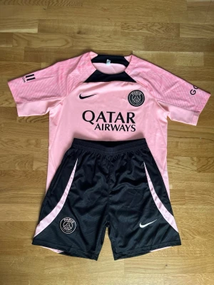 PSG rosa matchställ från Nike - Säljer ett rosa PSG matchställ med svart detalj från Nike. Tröjan har korta ärmar, svart Nike-logga och PSG-emblem på bröstet samt Qatar Airways-tryck. Shortsen är svarta med rosa detaljer och PSG-logga. Perfekt för fotbollsträning eller match.