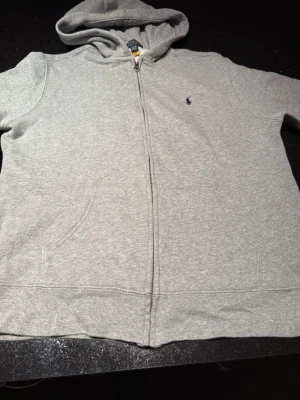 Grå zip hoodie från Polo Ralph Lauren - Snygg grå hoodie med dragkedja från Polo Ralph Lauren. Klassisk design med huva. Lite små slitningar på mudden i midjan annars bra skick
