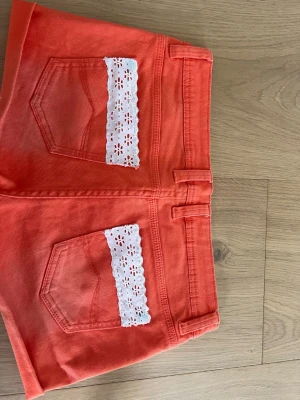 Korall shorts med spetsdetalj - Snygga korall shorts med vita spetsdetaljer på bakfickorna. Klassisk femficksmodell med bälteshällor och knappstängning. Perfekta för varma dagar och ger en färgstark touch till din outfit. Står att dom är storlek M/38 men skulle säga att dom är en blandning av m och s.