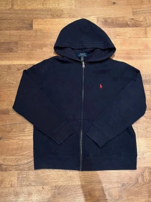 Marinblå hoodie från Polo Ralph Lauren - Klassisk Marinblå hoodie med dragkedja från Polo Ralph Lauren. Den har huva, två fickor framtill och den ikoniska röda loggan broderad på bröstet. Perfekt för en avslappnad och stilren look. Tillverkad i mjukt material för extra komfort.