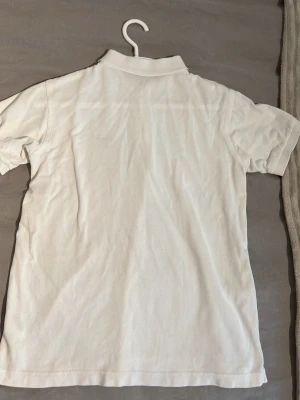 Vit pikétröja från Gant - Klassisk vit pikétröja från Gant med korta ärmar och krage. Tröjan har två knappar framtill och en liten broderad Gant-logga på bröstet. Tillverkad i mjuk bomull som känns skön mot huden. Perfekt till jeans eller shorts. Stolek saknas men den passar 12-13 år storlek 152