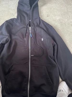 Svart zip hoodie från Polo Ralph Lauren - Säljer en svart hoodie med dragkedja från Polo Ralph Lauren. Klassisk design med huva och dragsko, två fickor framtill och den ikoniska vita loggan broderad på bröstet. Perfekt för en chill och stilren look.