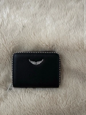 Svart plånbok med silverdetaljer - Snygg svart plånbok i ett stilrent format med silverfärgade nitar längs kanterna och ett emblem med vingar framtill. Plånboken har dragkedja runtom och är tillverkad i ett material som liknar skinn. Perfekt för dig som gillar en edgy och modern look.