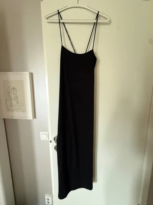 Svart långklänning - En stilren svart långklänning med tunna axelband och rak passform. Klänningen har minimalistisk design och är perfekt för dig som gillar en enkel men elegant look. Ä
