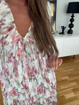 Blommig kortklänning med v-ringning - Säljer en luftig kortklänning med v-ringning och långa ärmar. Klänningen har ett romantiskt blommönster i rosa, grönt och vitt på plisserat tyg. Perfekt för sommarens alla tillfällen och ger en feminin och trendig look.