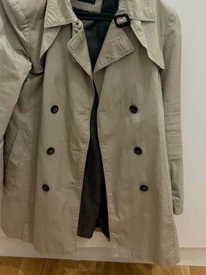 Beige trenchcoat med dubbelknäppning - Stilren beige trenchcoat med klassisk dubbelknäppning och bred krage.  Fodrad insida och raka ärmar ger en tidlös look. Perfekt för dig som vill ha en snygg och enkel trenchcoat till vardags.