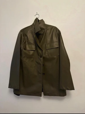 Olivgrön skinnjacka med fickor - Stilren olivgrön skinnjacka med rak passform och två bröstfickor med lock. Jackan har knäppning framtill och en hög krage som kan vikas upp för extra edge. Perfekt för dig som vill ha en cool och tidlös look.