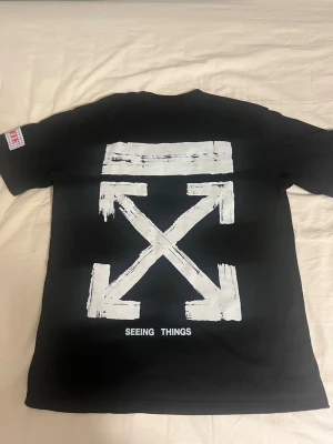 Svart Off-White t-shirt med tryck - Säljer en svart t-shirt från Off-White med ikoniskt vitt pilar-tryck på ryggen och texten 'SEEING THINGS'. Framtill står det 'OFF' och två vita detaljer längst ner. Klassisk passform och korta ärmar, tillverkad i mjuk bomull.