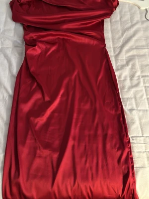 Röd långklänning i satin - Elegant röd långklänning i glansigt satinliknande material. Klänningen har en axelbandslös design med draperad överdel och slits baktill för extra rörelsefrihet. Perfekt för speciella tillfällen när du vill sticka ut.