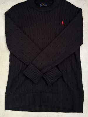 Svart kabelstickad tröja Polo Ralph Lauren - Svart kabelstickad tröja från Polo Ralph Lauren med rund halsringning och röd broderad logga på bröstet. Tröjan har ribbade muddar vid ärmslut och nederkant, och är tillverkad i ett mjukt stickat material.Priset kan diskuteras 🍾