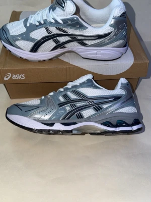 Asics vita och grå sneakers - Snygga Asics sneakers i vitt och grått med svarta detaljer. Ovandelen är i mesh och syntetmaterial för bra andningsförmåga och komfort. Klassisk Asics-logga på sidan och stabil sula med dämpning. Perfekta för en sportig och trendig look.