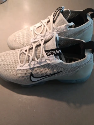 Nike Air Vapormax vita sneakers - Nike Air Vapormax sneakers i vitt med stickad mesh-överdel och svart Swoosh på sidan. Skorna har vita snören, svart detalj på plösen med 'VAPORMAX'-text och genomskinlig sula. Perfekta för dig som gillar sportig och modern stil. Size 39 fast passar även 42, 41, 40. Bara använd 2 gånger🤗. Köpte dom från England för  90 pund.